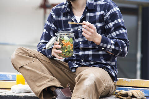 Close-up of construction worker holding salad in jar while sitting outdoors at site stock photo