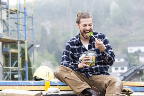 Construction worker eating salad while sitting outdoors at construction site - MJFKF00407