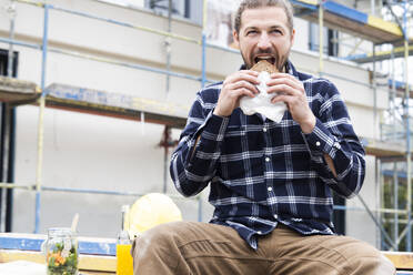 Construction worker eating bread while sitting against building at construction site - MJFKF00408