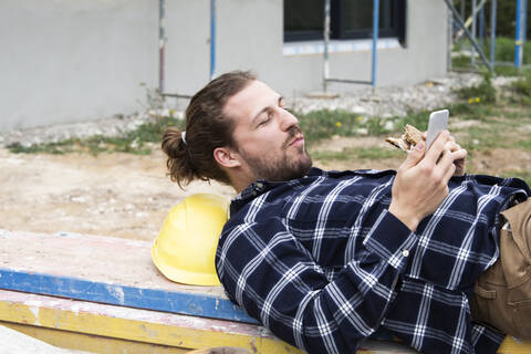 Bauarbeiter benutzt sein Smartphone, während er auf dem Holz liegt, lizenzfreies Stockfoto