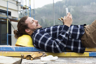 Construction worker using smart phone while eating food on wood at construction site - MJFKF00411