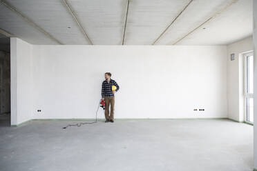 Construction worker holding work tool and helmet while standing in empty house - MJFKF00414