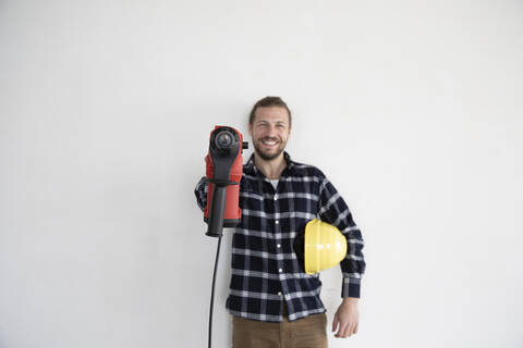 Smiling male worker holding work tool and helmet while standing against wall in house stock photo