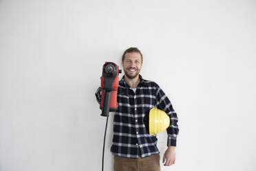 Lächelnder männlicher Arbeiter, der ein Arbeitswerkzeug und einen Helm hält, während er an der Wand eines Hauses steht - MJFKF00416
