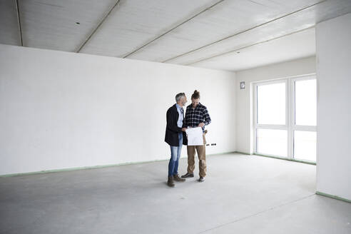 Architect and construction worker discussing blueprint while standing in empty house - MJFKF00417