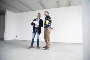 Architect and construction worker looking away while standing against wall in empty house - MJFKF00419