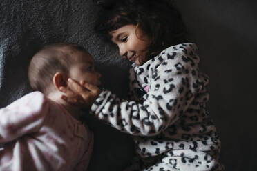 Mädchen berührt die Wangen ihrer kleinen Schwester, während sie auf dem Bett liegt - GEMF03920