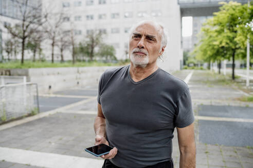 Älterer Mann hält Smartphone und schaut weg, während er auf einem Fußweg in der Stadt steht - MEUF01160