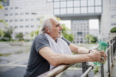 Senior man holding water bottle listening music while standing by railing - MEUF01167