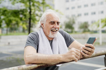 Nahaufnahme eines fröhlichen älteren Mannes, der sein Smartphone benutzt, während er an einem Geländer steht - MEUF01170