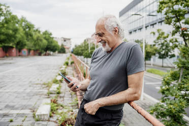 Lächelnder älterer Mann, der ein Smartphone benutzt, während er an einem Geländer in der Stadt steht - MEUF01190