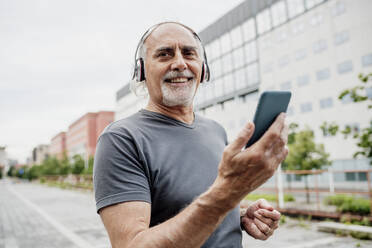 Smiling senior man wearing headphones using smart phone while standing in city - MEUF01192