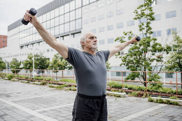 Serious senior man with arms outstretched holding dumbbells while standing on footpath - MEUF01193