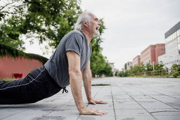 Senior man with mouth open doing push-ups on footpath in city - MEUF01200