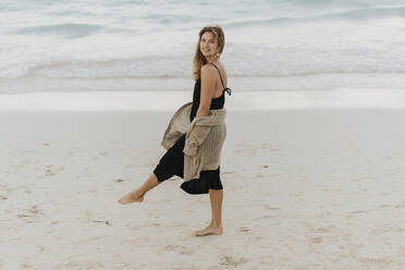 Lächelnde erwachsene Frau in schwarzem Kleid, die am Strand spazieren geht - SMSF00013