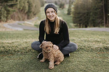 Smiling woman with poodle sitting on grassy land in park - SMSF00027