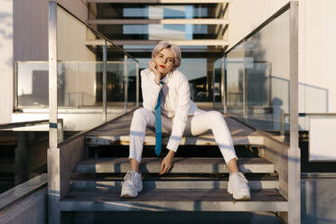 Young woman wearing white suit sitting on steps against modern building - TCEF00885