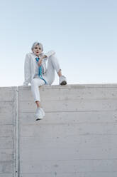 Junge Frau im weißen Anzug sitzt auf einer Stützmauer gegen den klaren Himmel - TCEF00890