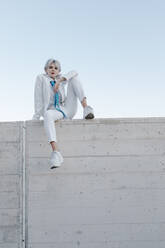 Young woman wearing white suit sitting on retaining wall against clear sky - TCEF00890