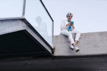 Junge Frau im weißen Anzug, die ihr Smartphone benutzt, während sie auf einer Stützmauer in der Stadt sitzt - TCEF00898