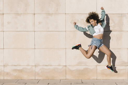 Excited young woman jumping against wall during sunny day - XLGF00307