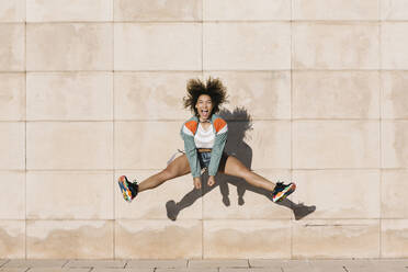 Excited young woman screaming while jumping against wall during sunny day - XLGF00308