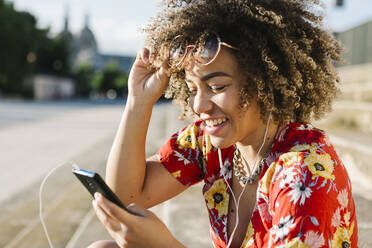 Happy trendy woman using smart phone while sitting on steps in city - XLGF00318