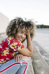 Glückliche junge Frau sitzt mit der Hand im Haar auf einer Treppe an einem sonnigen Tag - XLGF00323