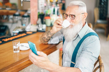 Bärtiger älterer Mann trinkt Kaffee, während er sein Smartphone im Café benutzt - EYF09498