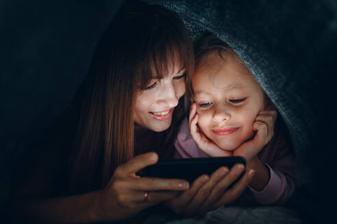 Lächelnde Mutter und Tochter schauen auf Handy unter der Bettdecke - EYF09521