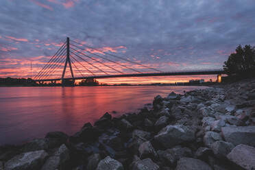 Deutschland, Nordrhein-Westfalen, Wesel, Felsiges Rheinufer bei dramatischem Sonnenuntergang mit Niederrheinbrücke Wesel im Hintergrund - KEBF01565