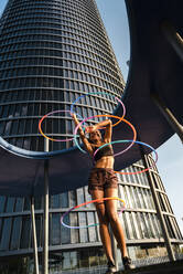 Fröhliche sportliche Frau, die mit Plastikreifen vor einem modernen Bürogebäude trainiert - JMPF00106