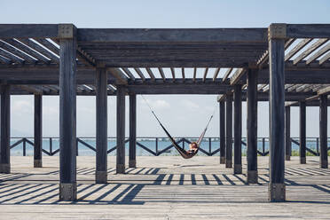 Woman lying in hammock hanging from metallic structure on boardwalk during sunny day - OCMF01418