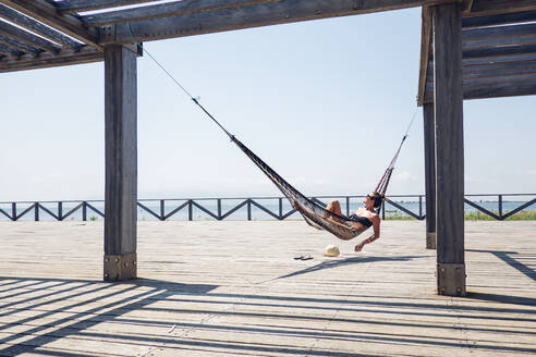 Entspannte Frau, die in einer Hängematte liegt, die an einer Metallkonstruktion an der Strandpromenade hängt, während eines sonnigen Tages - OCMF01419
