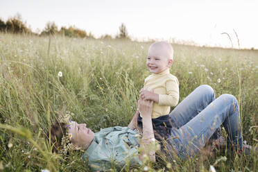 Happy boy sitting on mother lying in grassy field during sunny day - EYAF01214