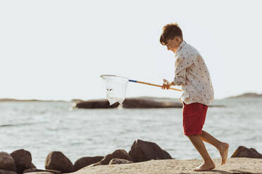 Smiling boy with fishing net walking over rock formation against clear sky during sunny day - MASF18435