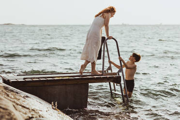 Seitenansicht einer sprechenden Frau, während ihr Sohn auf einer Leiter im Meer steht - MASF18449