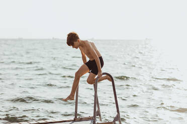 Playful shirtless boy balancing on ladder against sea during sunny day - MASF18450