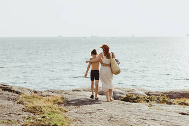 Rear view of woman with son walking against sea during sunny day - MASF18459