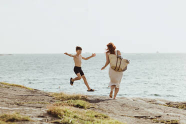 Rear view of woman with bag while son jumping against sea during sunny day - MASF18460