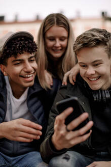 Glückliche Teenager, die in der Stadt sitzend soziale Medien auf ihrem Mobiltelefon nutzen - MASF18500