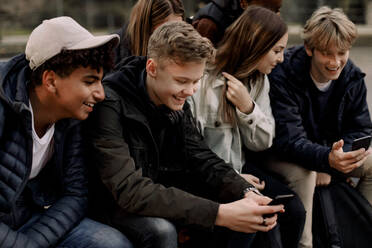 Lächelnde Mädchen und Jungen im Teenageralter, die im Park sitzen und Mobiltelefone benutzen - MASF18505
