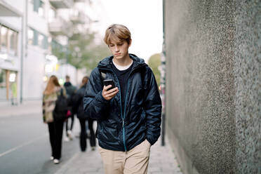 Teenage boy with blond hair using smart phone while walking on sidewalk against wall in city - MASF18508
