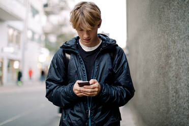 Teenage boy using mobile phone while walking on sidewalk against wall in city - MASF18509