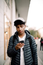 Teenage boy wearing cap using smart phone while walking on footpath in city - MASF18512