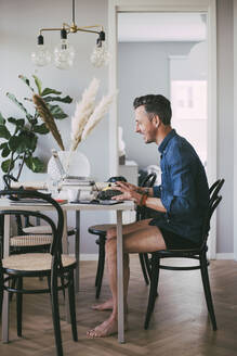 Happy man sitting at table in underwear working at home having a video call - MASF18531