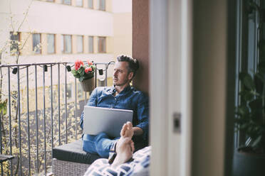 Mann sitzt auf Balkon mit Laptop und schaut hinaus - MASF18547