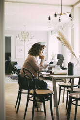 Woman sitting at table at home talking to coworkers in online meeting - MASF18593