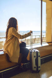 Businesswoman holding wheeled luggage while sitting on window sill at airport departure area - EHF00380