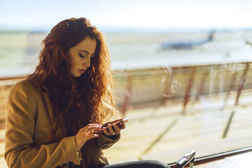 Businesswoman using smart phone by window at airport departure area - EHF00383